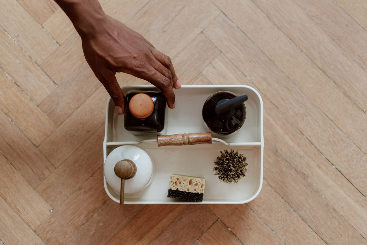A white caddy of wooden cleaning brushes, soap and cloths on a wooden floor