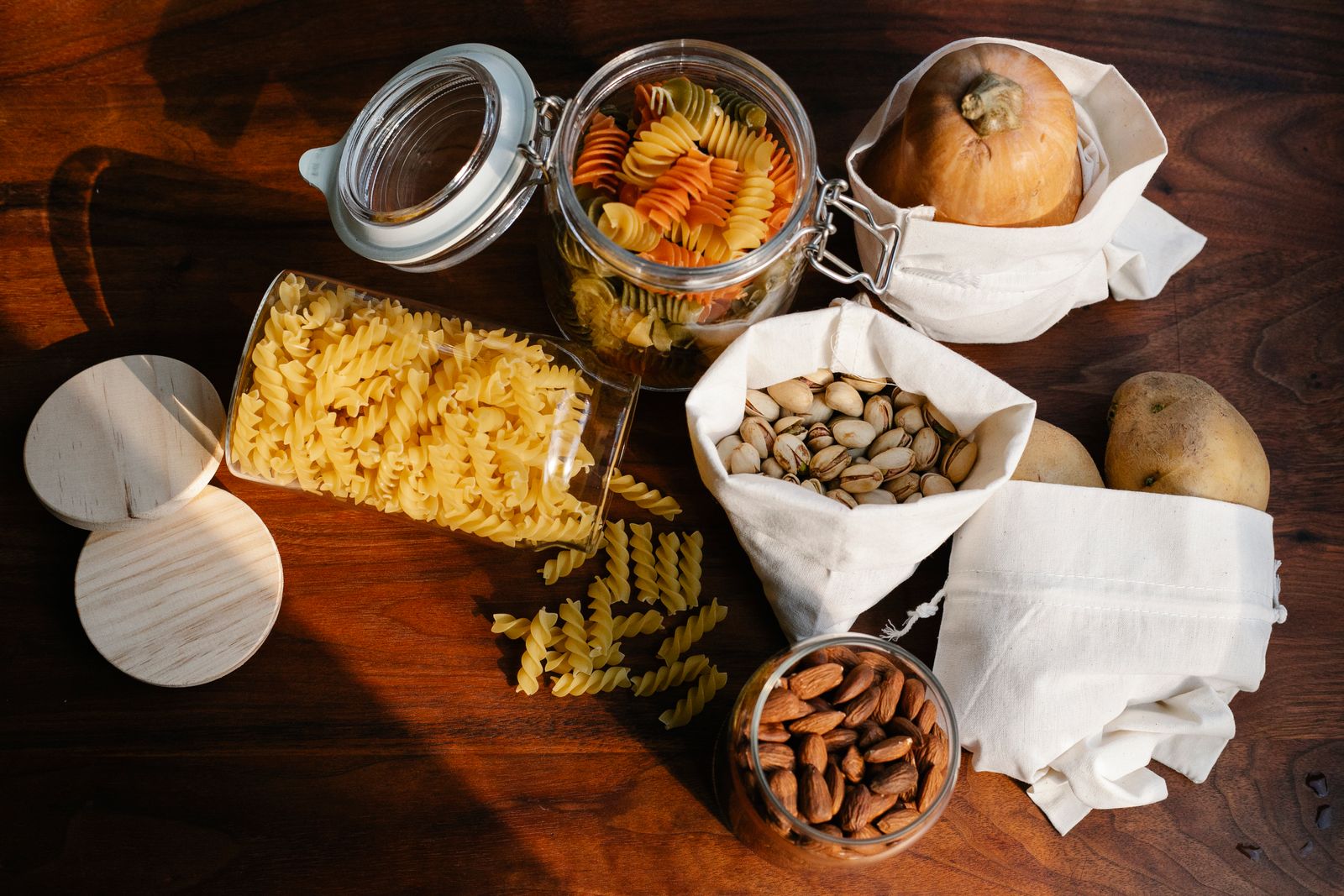Glass jars of pasta and nuts beside cotton drawstring produce bags on a wooden kitchen table