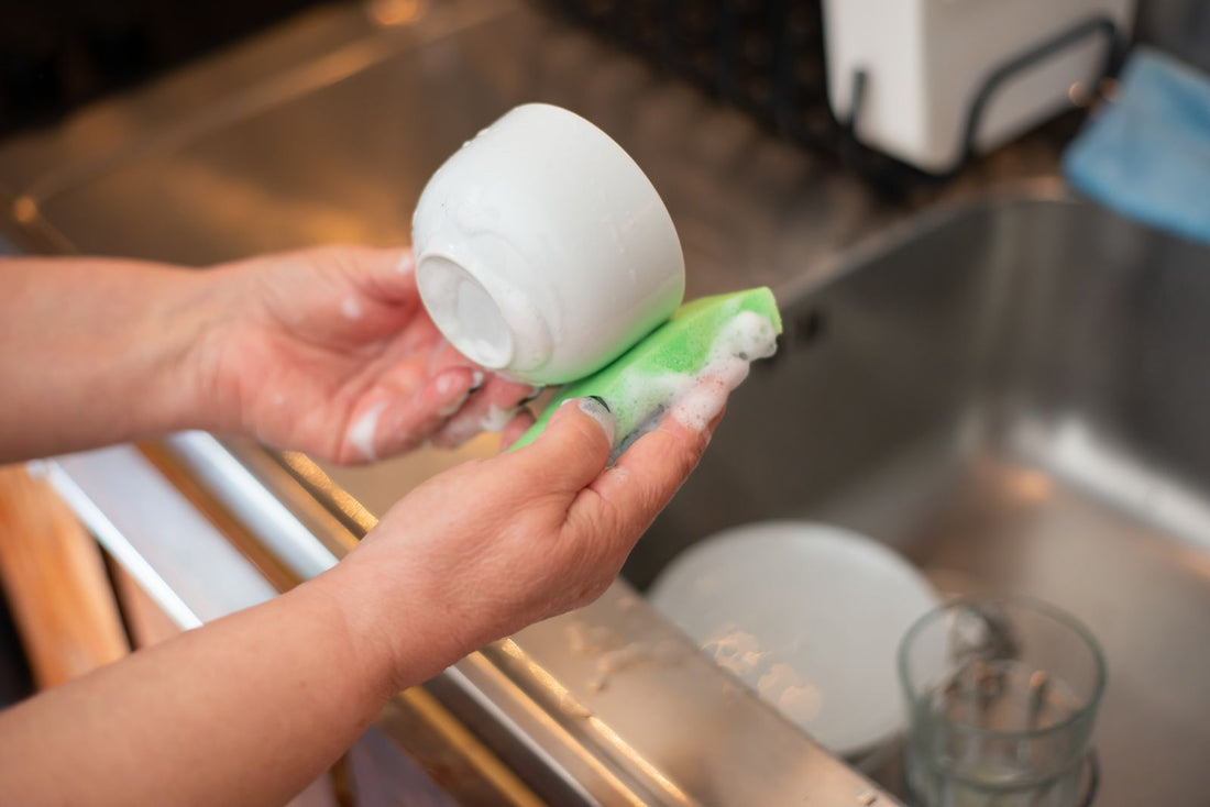 Close up of hands washing a white cup with a sponge and soap suds over a kitchen sink